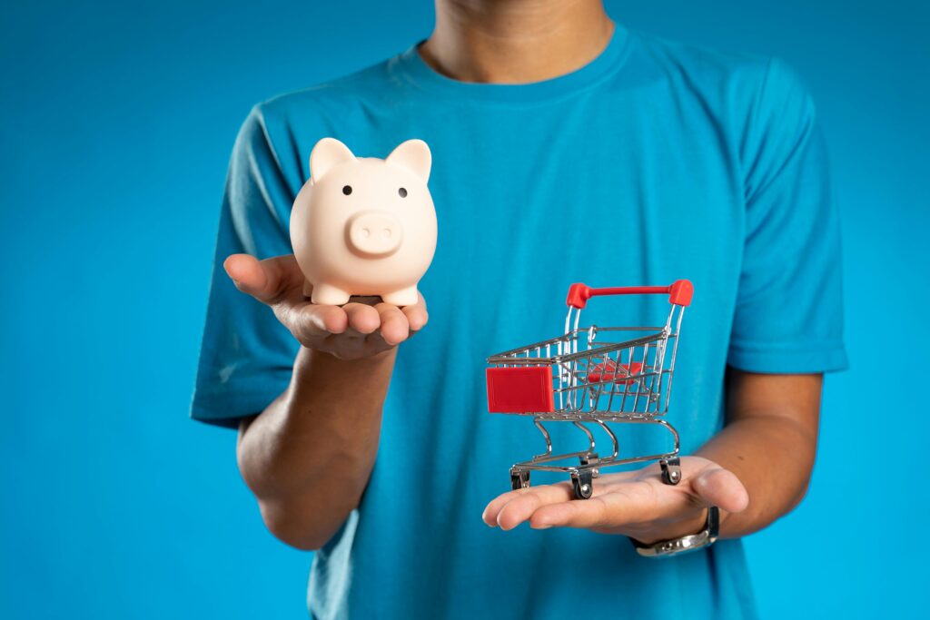 pexels photo 12357425 12357425 Man in blue shirt holding piggy bank and mini shopping cart, symbolizing savings versus spending.